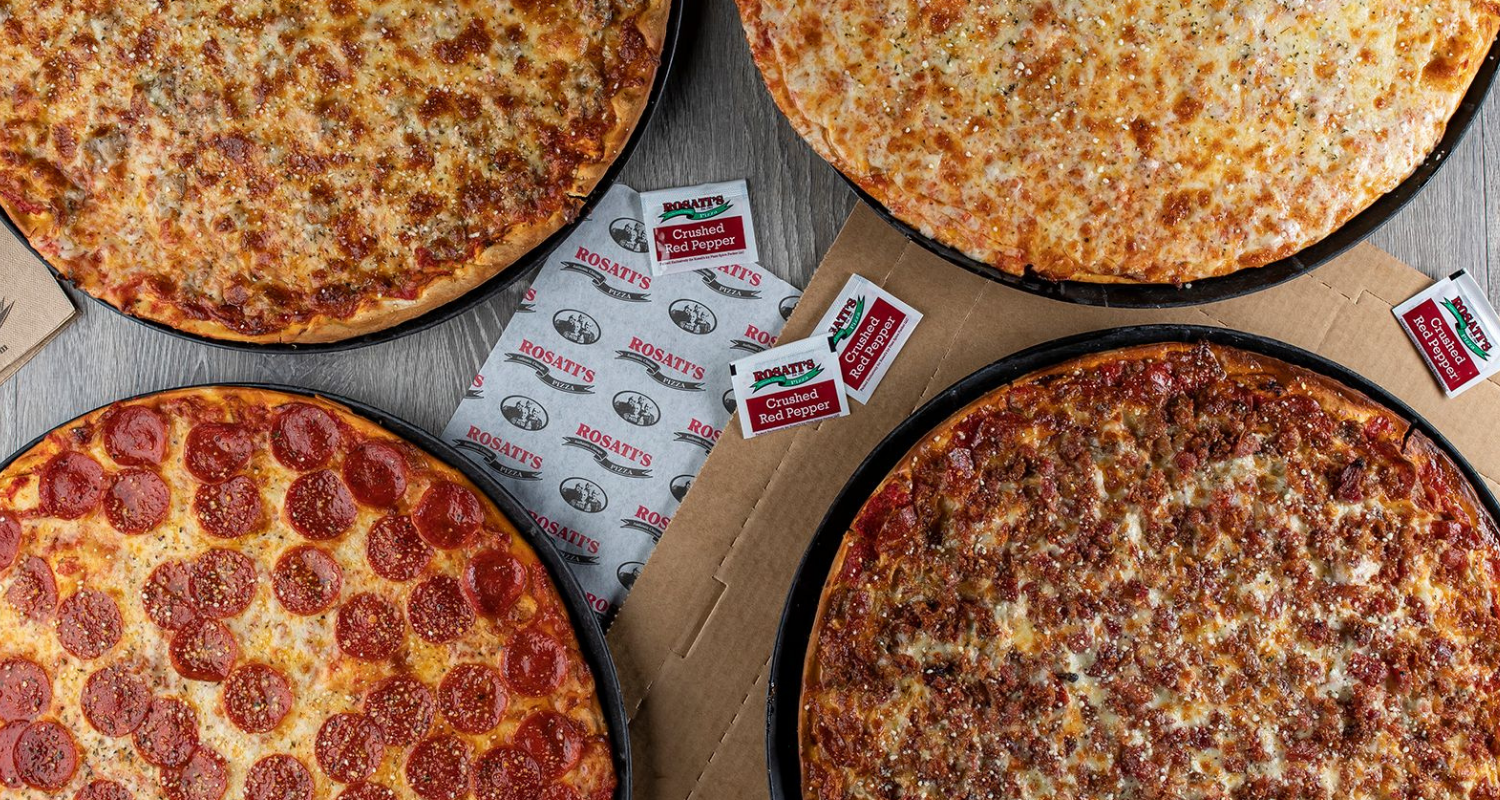Four pizzas on a table with a bunch of condiment packets in the middle.