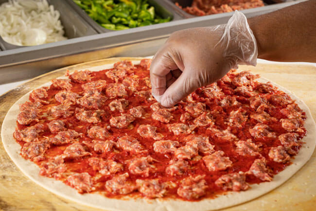Picture of a gloved hand putting toppings on a pizza with marinara sauce